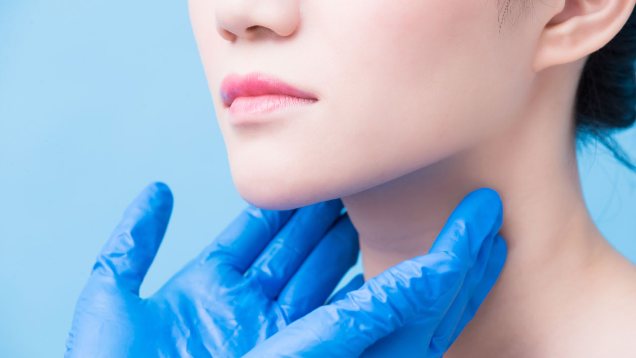 foto de parte do rosto e pescoço de uma mulher, com duas mãos vestidas com luvas azuis apalpando sua tireoide